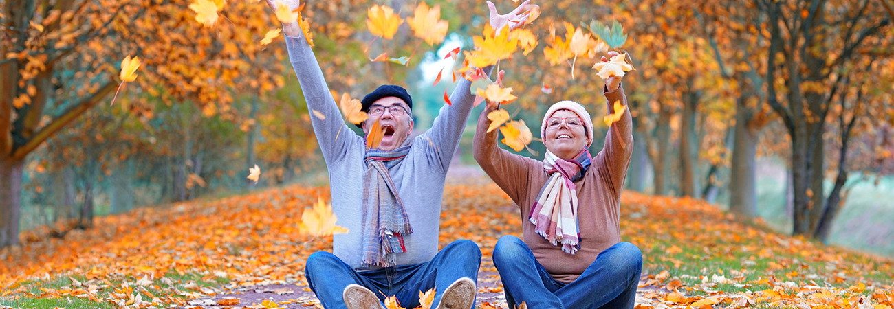 Zwei fröhlich aussehende Personen sitzen auf einer Straße und schmeißen Herbstlaub in die Luft.