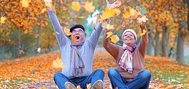 Zwei fröhlich aussehende Personen sitzen auf der Straße und werfen Herbstlaub in die Luft.